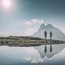 Due escursionisti con bastoni al lago di fronte a un paesaggio montano con cielo soleggiato